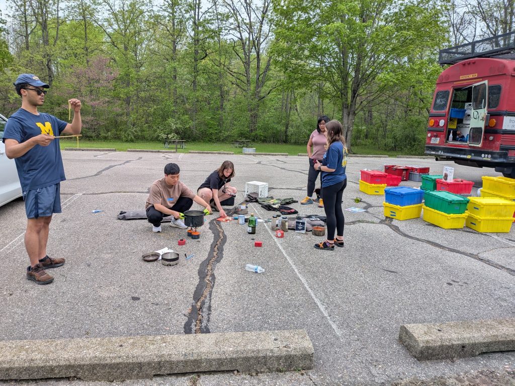 An instructor demonstrates camp stove use beside the Get Outside Bus with gear bins laid out
