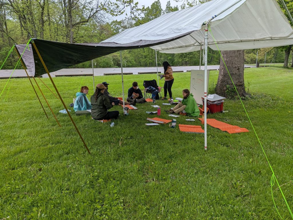 Course participants seated under a tarp shelter for an outdoor classroom session